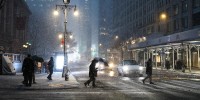 Pedestrians carrying umbrellas navigate heavy snowfall on the streets of Manhattan at night.