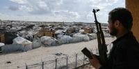 A person holding a gun looks towards a village of tents outside