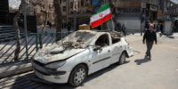 A man walks past an Iranian flag fluttering above the wreckage of a car.
