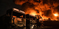 Charred vehicles seen outside near plumes of smoke and fire in the distance