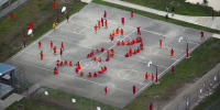 An aerial view of dozens of detainees wearing orange prison uniforms on a basketball court, arranging their bodies to spell SOS.