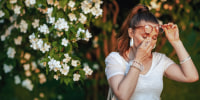 Summer time. unhappy elegant female in white shirt with handkerchief and eyeglasses has an allergy attack near flowering tree.