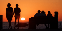 People watch the sunset at Aliso Beach, their silhouettes are seen as they face the water and horizon, orange sky