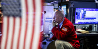 A person sits at a work desk with various monitors, an American flag covers half of the photo in the foreground