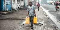 A man carries two yellow water jugs outside on the sidewalk