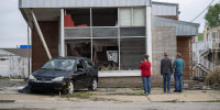 People stand outside of a damaged building with a damaged car parked out front