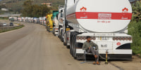 A man sits on a folding chair in front of a long line of tanker trucks parked alongside a dusty highway. 