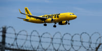 A yellow Spirit Airlines planes can be seen flying above a barbed wire fence in the foreground