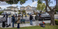 A line of reporters with television cameras stand across the street from a row of two story residential homes on a tree-lined street.