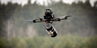 A wired drone hovering in front of a distant treeline.
