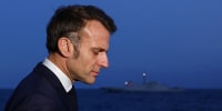Emmanuel Macron appears in profile view in front of a battleship floating in the sea at dusk.