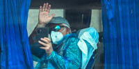 A person wearing sunglasses, a face mask, and a blue plastic hazard suit waves from a bus window while holding their phone up