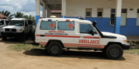 A white 4x4 with red lettering reading AMBULANCE parked on a dirt road outside a one-story concrete building.