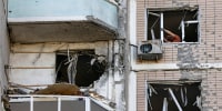 Residents clear broken windows in a damaged apartment building, as seen from the outside of the building