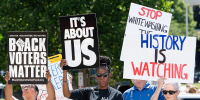People hold signs that say "Black Voter Matter", "It's About Us", and "History Is Watching" while standing outside