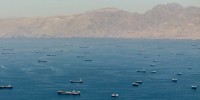 An aerial view of dozens of large ships anchored in a waterway in front of tan mountains.