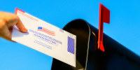 Woman's hand placing a mail-in election ballot in a mailbox against a blue sky