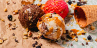 Chocolate, strawberry and vanilla ice cream scoops with caramel drizzle, scattered nuts and blueberries on parchment paper and a mint patterned surface.