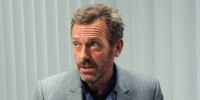 Hugh Laurie sits inside of a doctor's office next to medical supplies as he speaks to a patient sitting in front of him