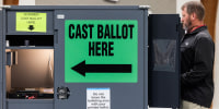 James Couch votes at a voting booth inside of a room, a large green sign that reads "cast ballot here" in black text is on the side of the voting booth