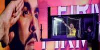 Abelardo de la Espriella raises a fist into the air while speaking into a microphone from inside an elevated, bulletproof glass cube.