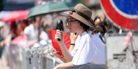 A woman wearing a hat holds an electric fan to cool her face while standing behind a metal barricade