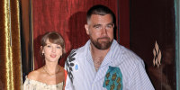 Travis Kelce, looking toward the camera, holds Taylor Swift's hand as they exit a building to the street.