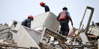 Workers wearing red helmets look around while walking up a pile of rubble