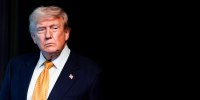 Donald Trump stands and looks off to the side, against a black background that his suit blends into