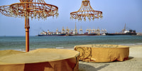 Two tables with chandeliers on a beach in front of large container ships floating in the distance.