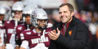 Seth Bodnar stands and claps while smiling near blurred football players outside on the field