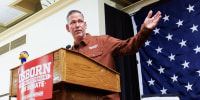 Dan Osborn speaks at a podium and extends his hand outwards, there is a red poster on the front of the podium that reads in white text: "Osborn, lifelong independent, for senate. To fix DC, send a mechanic."