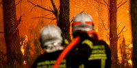 Fire and sparks can be seen in the woods, the backs of two firefighters can be seen