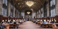 A large stone room with a vaulted ceiling and chandelier and long rows of wooden desks.