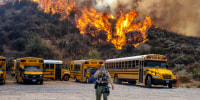 A brush fire is seen in the trees behind a row of parked school buses in a parking lot as a cop walks in the foreground speaking