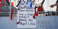 A sign that reads "Say His Name Edwin Lopez Cornejo Died in ICE/GEO custody at Delaney Hall August 1, 2026" hangs on a chainlink fence outside amongst tied ribbons and an El Salvadorian flag draped above it. Written in marker on the concrete wall below the chainlink fence, next to the poster, reads "Free Them"
