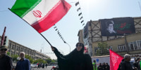 Image: A woman waves an Iranian flag. A banner showing a portrait of the late Supreme Leader Ayatollah Ali Khamenei is seen hanging on a building wall in the background.
