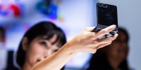 A woman holds a Samsung Galaxy Z Flip 8 up into the air while looking into it