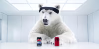 A polar bear looks at a can of Pepsi, left, and a can of Coca-Cola while seated in an all-white room at a table