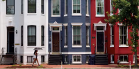 A woman with an umbrella walks past red, white and blue painted row houses.