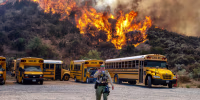 A brush fire is seen in the trees behind a row of parked school buses in a parking lot as a cop walks in the foreground speaking