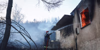 A firefighter sprays a hose into the window of a house on fire, in a charred forest environment