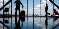 Silhouettes of people standing in an airport near large windows with a view of the sky and tarmac, reflections can be seen on the floor