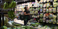 A woman reaches toward the top shelf of a refrigerated grocery store shelf.