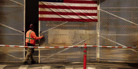 A construction worker wearing an orange vest and hardhat stands beneath a large American flag on a fence.