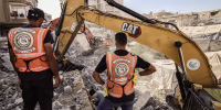 Two men wearing orange vests stand in front of two diggers in a field of debris.