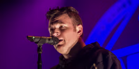 Chad Gilbert singing into a microphone under stage lighting.