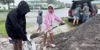 A young girl shovels wet sand into a sandbag in the rain.