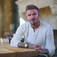 David Beckham in a white henley T-shirt with buttons and a wristwatch sits at a table and looks serious. 