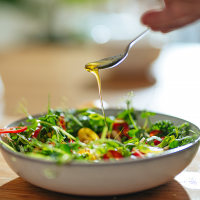 Hand pouring a teaspoon olive oil into a fresh salad with micro green herbs at the kitchen table.